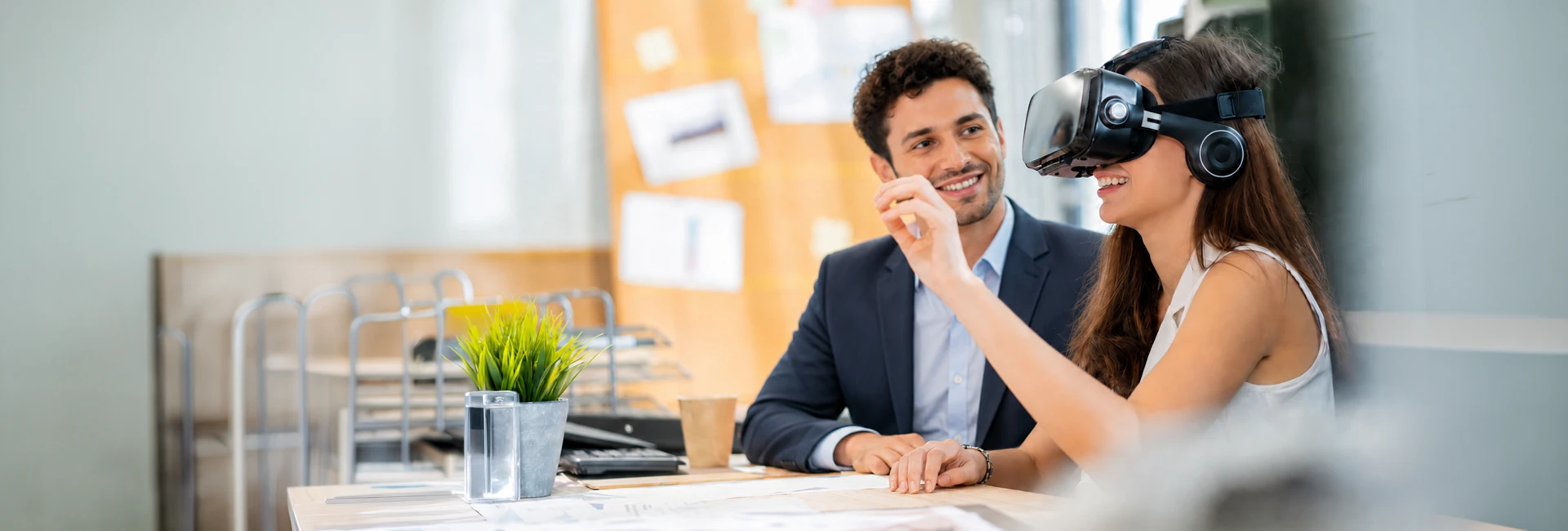 Two colleagues at a desk in a bright office, one wearing a virtual-reality headset