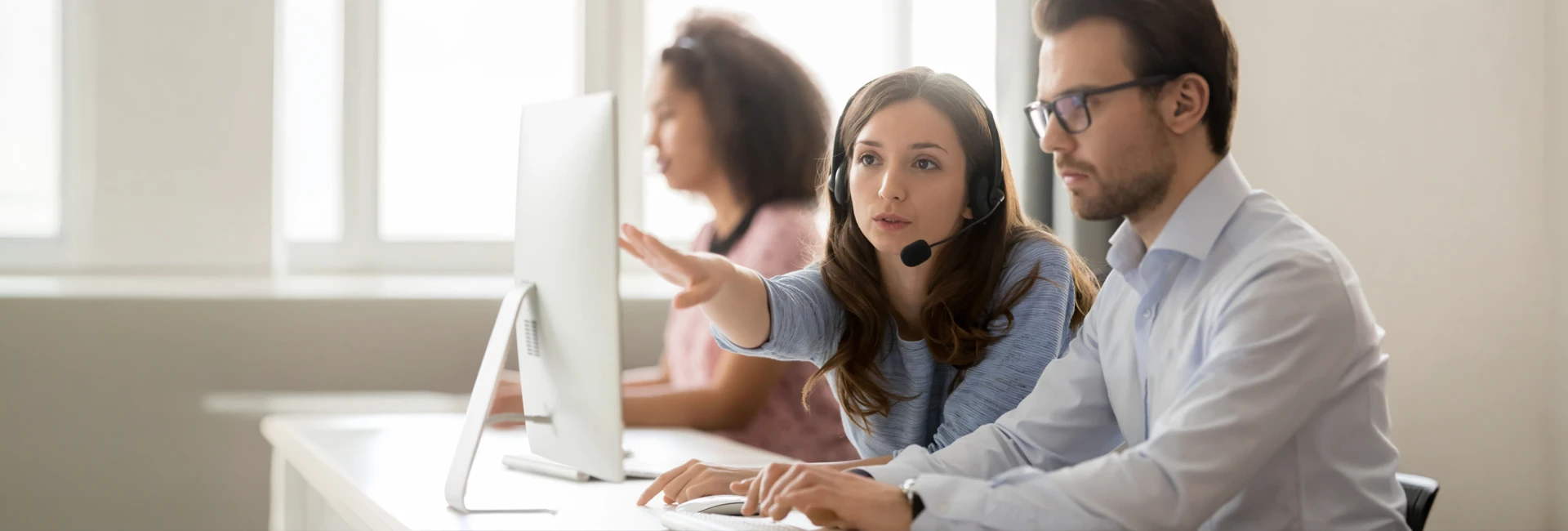 Colleagues at a desk in a bright office, one wearing a headset and pointing at a monitor