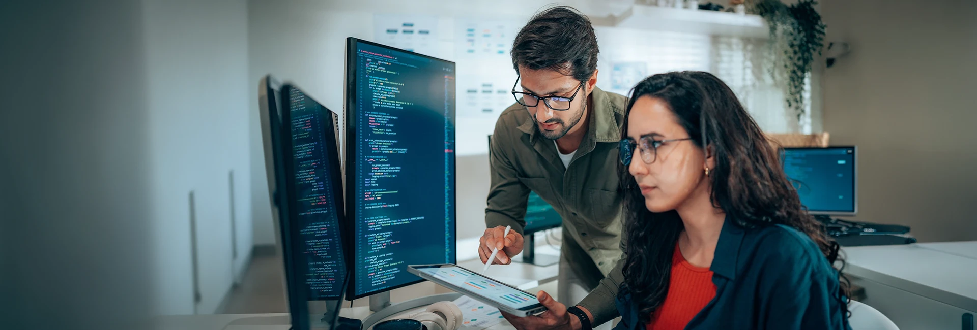 Two engineers reviewing code on a monitor while one points at a dashboard on a tablet